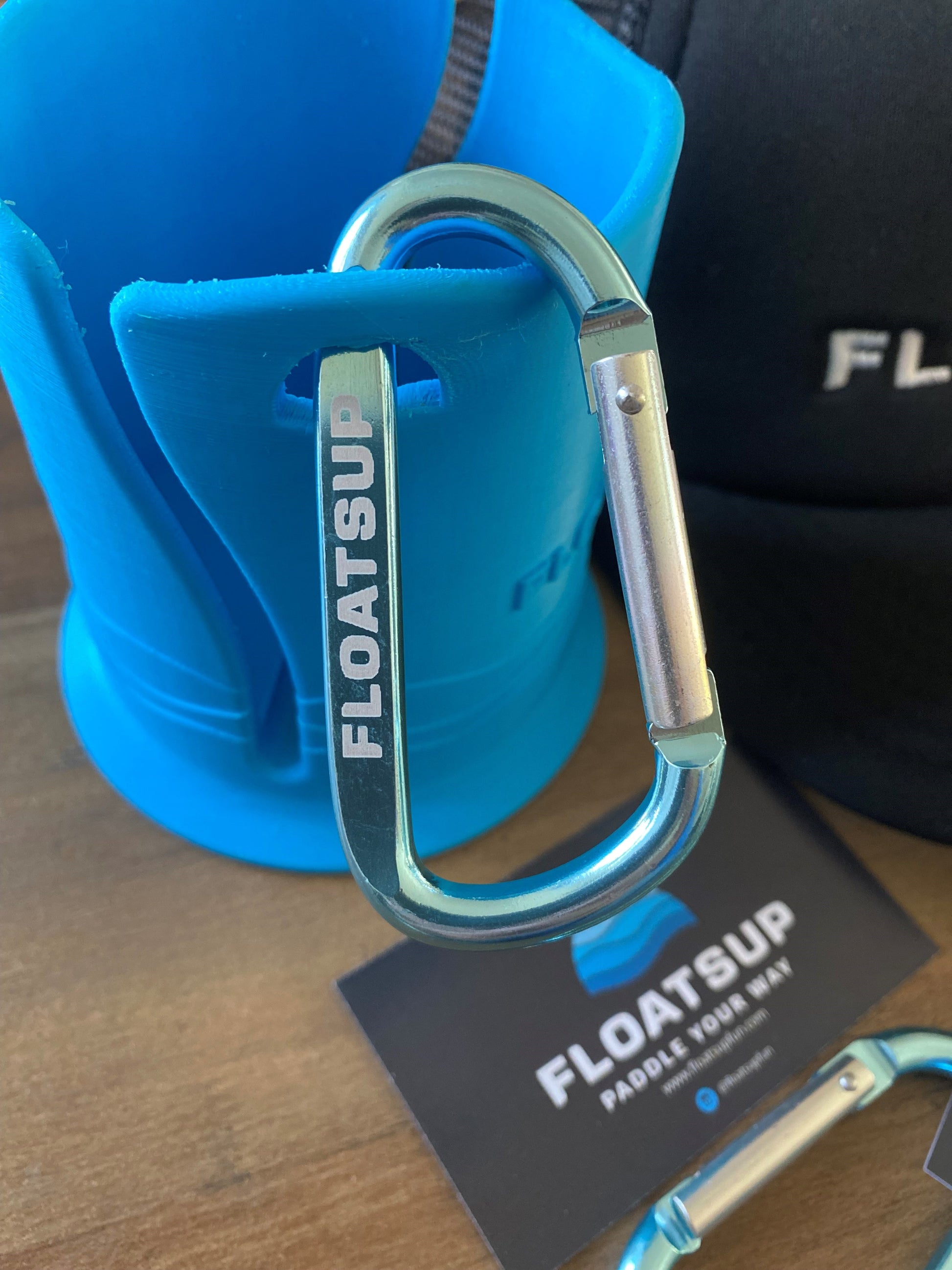 Floatsup carabiner in a light aqua color attached to a Floatsup Cup paddle board drink holder. Carabiner says "Floatsup" on it on one side and www.floatsupfun.com on the other.